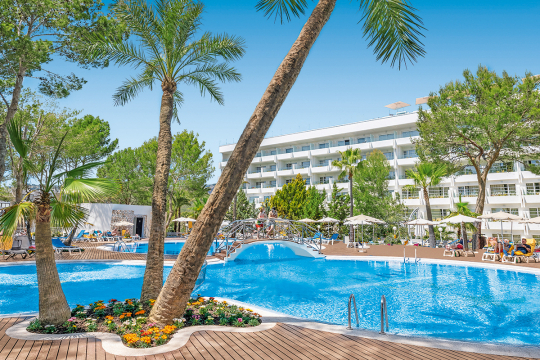 Poolanlage eines Hotels in ovaler Form mit Palmen, Liegen und im Hintergrund das wei&szlig;e Hotelgeb&auml;ude